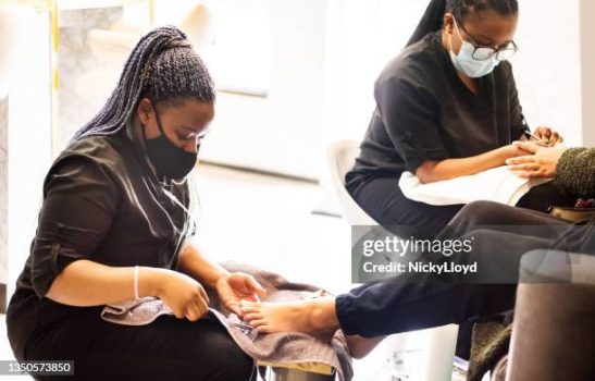 Two beauticians wearing face masks giving pedicure and manicure to a client in beauty salon