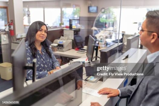 A female Hispanic bank teller smiles while helping a male customer at the bank counter.