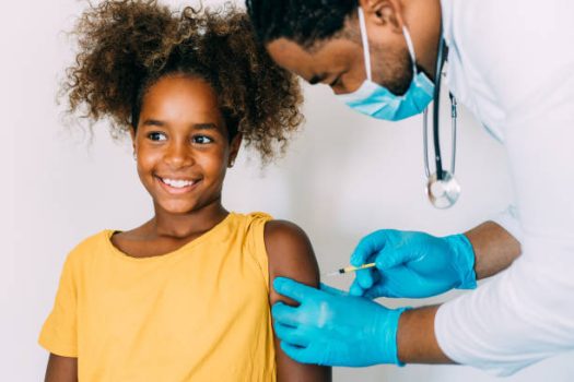 Male doctor wearing blue latex protective gloves injecting a child in her arm with a needle and syringe containing a dose of the COVID-19 vaccine.